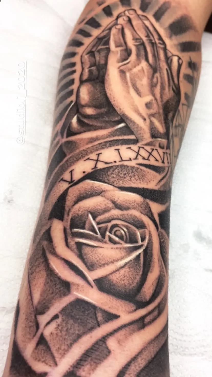 Tatuagem de mãos em oração com rosa e faixa romana no antebraço